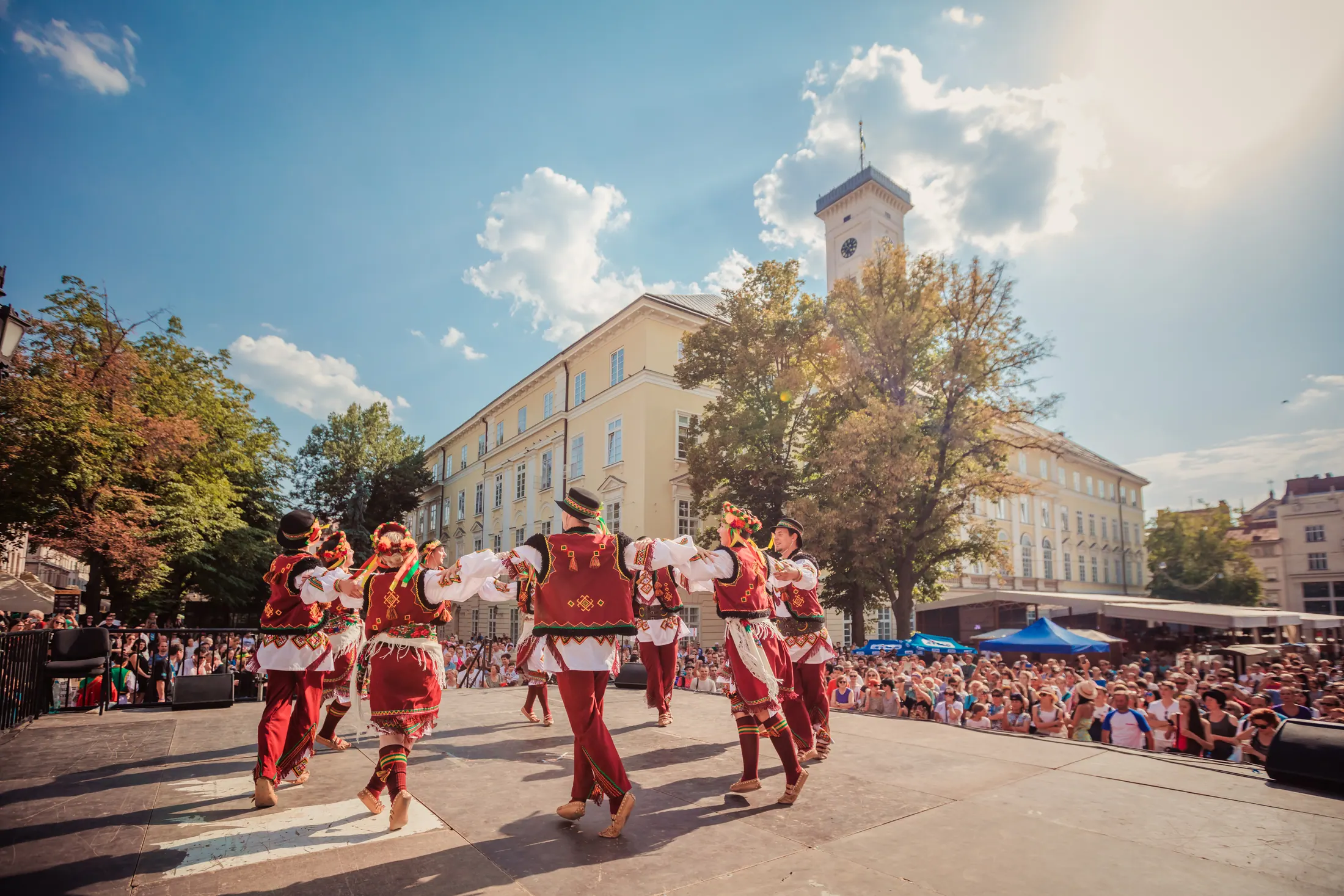 A circle of dancers in red and maroon traditional costume performing for a large crowd in front of Lviv's city hall